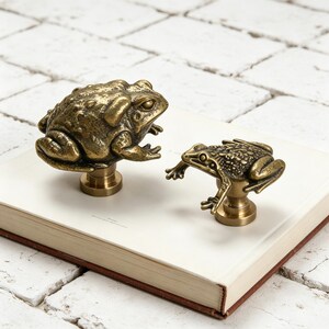 May include: Two brass frog-shaped cabinet knobs. One knob is larger and depicts a frog in a seated position, while the other is smaller and shows a frog in a crouching pose. The knobs are mounted on a white surface.