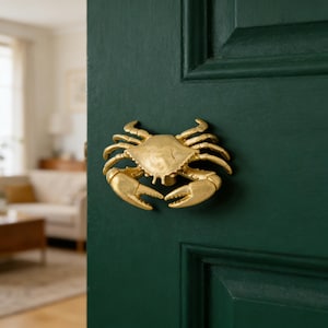 Könnte beinhalten: Ein goldfarbener Türklopfer in Form einer Krabbe, montiert an einer dunkelgrünen Tür. Der Klopfer ist eine detaillierte, dreidimensionale Skulptur einer Krabbe mit ausgestreckten Scheren. Die Tür ist in einem satten, tiefen Grün gestrichen.