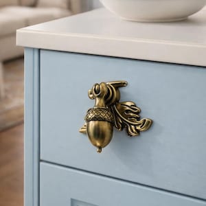 May include: A brass acorn-shaped cabinet pull with intricate leaf detailing. The hardware is mounted on a light blue cabinet door. A white countertop and a bowl of fruit are in the background.