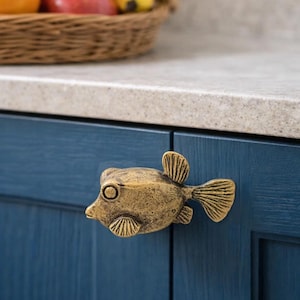 Könnte beinhalten: Ein messingfarbener, fischförmiger Schrankknauf ist an einer dunkelblauen Schranktür angebracht. Der Knauf hat detaillierte Flossen und ein rundes Auge. Ein Korb mit Obst steht auf der Arbeitsplatte über dem Schrank.