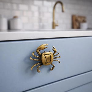 May include: A brass crab-shaped cabinet pull mounted on a light blue cabinet door. The crab has detailed claws, legs, and a textured body. The background shows a white countertop and a gold faucet.