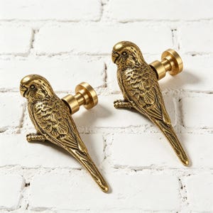 May include: Two brass bird-shaped cabinet knobs. The knobs are detailed with feathers and have a gold finish. The birds are perched on a round base with a screw post. The knobs are set against a white brick wall.