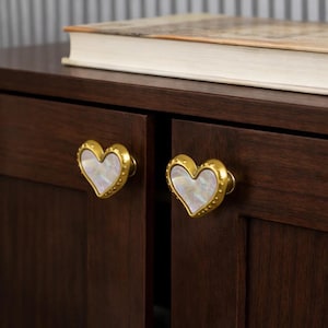 May include: Close-up of a dark brown cabinet with two heart-shaped drawer pulls. The pulls have a gold-colored trim with a pearl-like center. A book rests on top of the cabinet.
