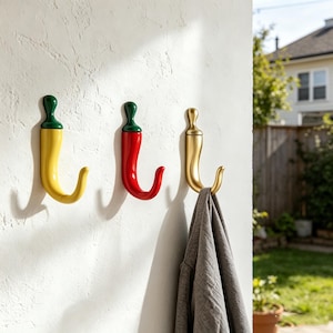 May include: Three decorative wall hooks shaped like chili peppers. One is yellow with a green stem, another is red with a green stem, and the third is gold. A gray garment hangs from the gold hook. The hooks are mounted on a white wall.