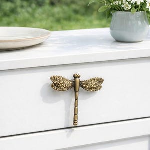 Peut inclure: Un bouton de tiroir décoratif en forme de libellule en laiton est centré sur une armoire blanche. La libellule a des ailes détaillées et un long corps segmenté. Un vase de fleurs blanches à cœur jaune est à droite.