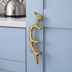 May include: A gold-colored, branch-shaped cabinet pull is mounted on a light blue cabinet door. The pull has a unique, organic design. A stainless steel kettle with a blue handle is visible in the background.