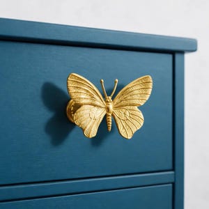 May include: A gold-colored butterfly-shaped drawer pull mounted on a dark teal blue cabinet. The butterfly has detailed wing patterns and a textured body. The cabinet has a simple, clean design with visible drawer lines.