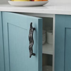 Puede incluir: Primer plano de una puerta de armario de cocina azul claro con un tirador negro en forma de hoja. La puerta del armario está ligeramente entreabierta, revelando estantes blancos y vajilla en el interior. Un cuenco de fruta está sobre la encimera.