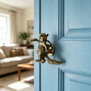 May include: A brass squirrel door handle is mounted on a light blue door. The squirrel is holding a flute and a bag. The door has a paneled design. The background shows a living room with a sofa and a coffee table.