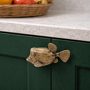 May include: A bronze-colored fish-shaped cabinet knob. The knob is mounted on a dark green cabinet door. The fish has detailed fins and a textured surface. A woven basket with produce is in the background.