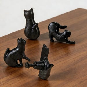 Puede incluir: Cuatro pomos de armario negros con forma de gato sobre una superficie de madera. Los pomos están en diferentes poses, incluyendo sentado, estirándose y uno con un tornillo adjunto. Los pomos están hechos de un material texturizado.