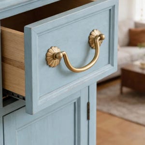 May include: A light blue cabinet with a drawer partially open, revealing a wooden interior. The drawer features a gold-colored handle with a decorative floral design. The cabinet has a distressed finish, giving it a vintage look.