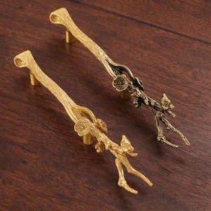 May include: Two decorative cabinet pulls with a branch-like design. One is gold-toned, the other is bronze. Both have floral accents. The pulls are made of metal and are set against a dark wood surface.