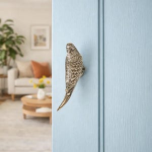 May include: A decorative brass bird-shaped cabinet pull mounted on a light blue cabinet door. The bird is detailed with etched feathers and a curved beak. The pull adds a touch of nature-inspired elegance to furniture.