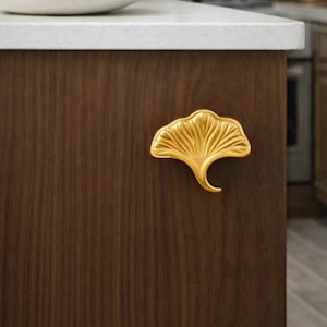 Può includere: Una maniglia dorata a forma di foglia di ginkgo è montata su un'anta di un mobile in legno marrone scuro. L'anta del mobile fa parte di un'isola della cucina. Un piano di lavoro bianco e una fruttiera sono visibili.