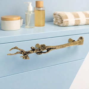 Puede incluir: Un tirador de cajón de color bronce en forma de rama de árbol con pequeñas flores. El tirador en forma de rama está montado en un mueble azul claro. Otros accesorios de baño son visibles en el fondo.