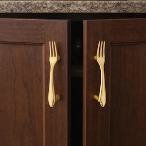May include: Close-up of two dark brown cabinet doors with gold-colored fork-shaped handles. A wooden bowl with fruit sits on the countertop above the cabinets. The handles add a unique touch to the kitchen.