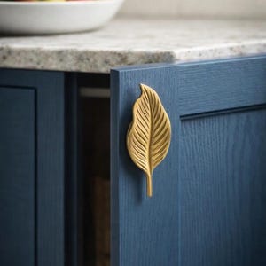 May include: A gold-colored leaf-shaped cabinet pull is mounted on a dark blue cabinet door. The cabinet door is slightly ajar, revealing the interior. A white bowl of fruit sits on the countertop in the background.
