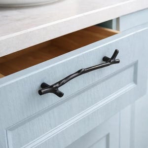 Könnte beinhalten: Schwarzer, astförmiger Schrankgriff an einer hellblauen Schranktür. Die Schublade ist geöffnet und gibt einen Einblick in das Holzinnere. Eine weiße Arbeitsplatte und ein Teller mit Obst sind im Hintergrund zu sehen. Diese Hardware verleiht eine natürliche, rustikale Note.