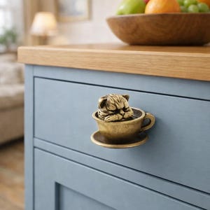 May include: A brass cabinet knob shaped like a cat in a teacup. The cat is curled up inside the cup, with its head resting on the rim. The knob is mounted on a light blue cabinet door.