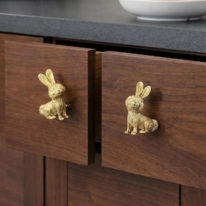 Puede incluir: Dos puertas de armario de madera con tiradores de cajón dorados en forma de conejito. Los tiradores están detallados con pelaje texturizado y poses sentadas. Las puertas del armario son de color marrón oscuro. Un cuenco de fruta es visible en el fondo.