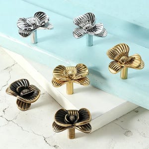 May include: A selection of flower-shaped cabinet knobs in silver, gold, and bronze. The knobs have detailed petal designs and are mounted on short stems. The hardware is displayed on a light blue and white surface.