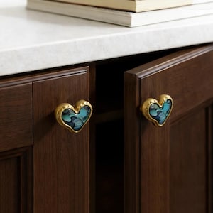 Può includere: Pomelli per armadi a forma di cuore con finitura dorata e design floreale blu e verde. I pomelli sono montati su ante di armadi marrone scuro. I pomelli aggiungono un tocco decorativo agli armadi della cucina o del bagno.