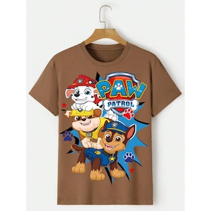 May include: A brown t-shirt featuring the Paw Patrol logo and three cartoon puppies. The puppies are wearing hats and uniforms in red, yellow, and blue. The Paw Patrol logo is in blue and red with a paw print.