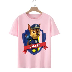 May include: Pink t-shirt featuring a graphic of Chase from Paw Patrol. The design includes Chase in his police uniform, a blue shield, and the name "CHASE" on a red banner. The shirt has a crew neck and short sleeves.