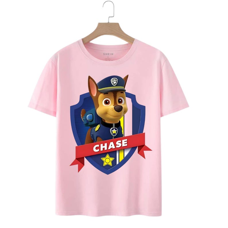 May include: Pink t-shirt featuring a graphic of Chase from Paw Patrol. The design includes Chase in his police uniform, a blue shield, and the name "CHASE" on a red banner. The shirt has a crew neck and short sleeves.