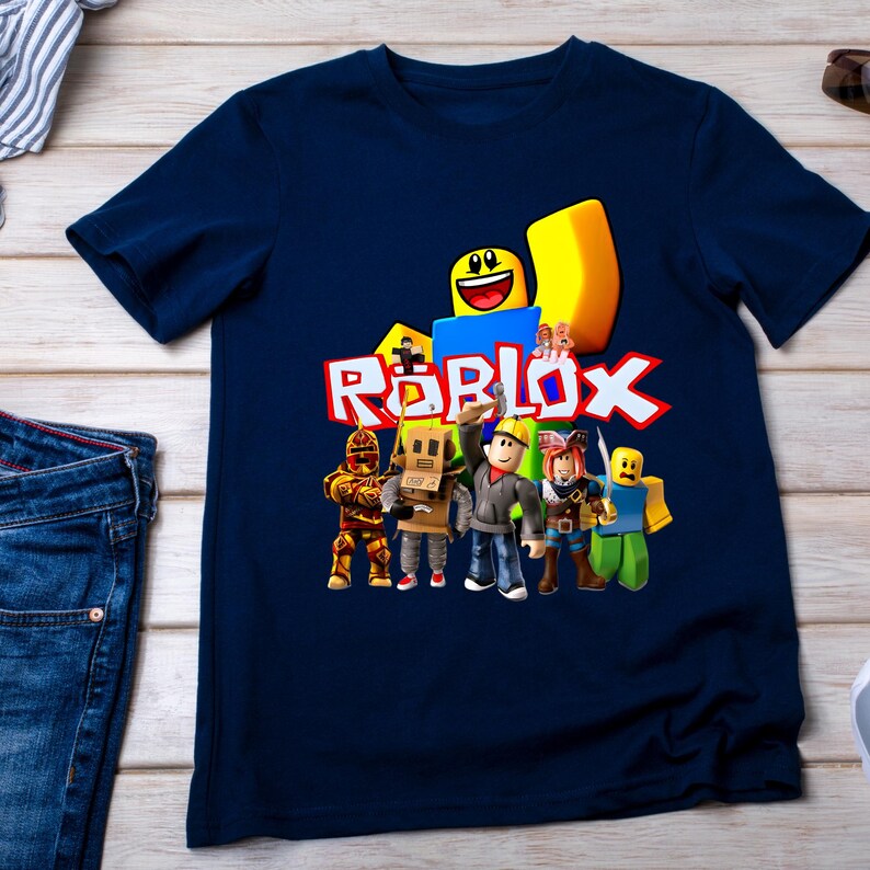 May include: Navy blue t-shirt with the Roblox logo and character illustrations. The design includes a yellow block, a smiling face, and figures in various outfits. The word "ROBLOX" is in red and white.