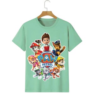 May include: A light green t-shirt featuring the Paw Patrol characters. The design includes the Paw Patrol logo and images of the cartoon characters. The shirt is hanging on a wooden hanger.