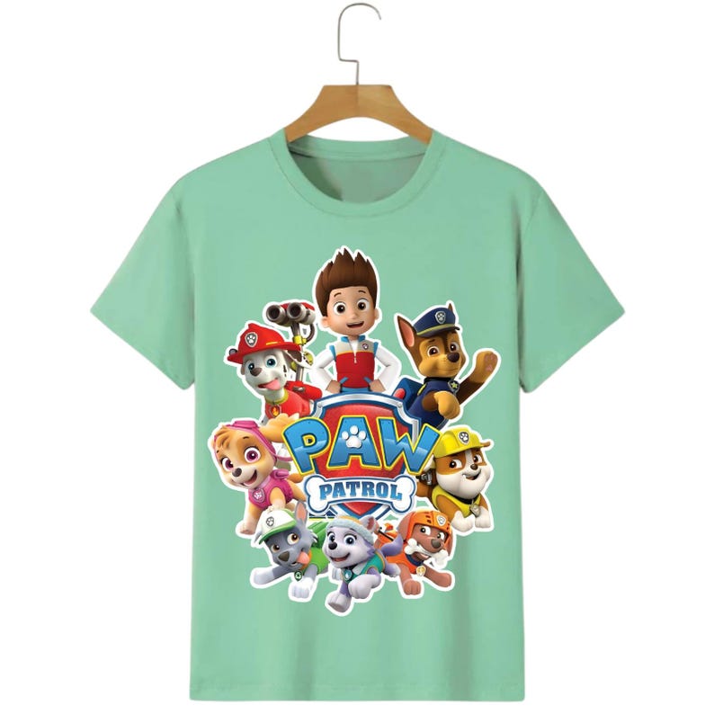 May include: A light green t-shirt featuring the Paw Patrol characters. The design includes the Paw Patrol logo and images of the cartoon characters. The shirt is hanging on a wooden hanger.