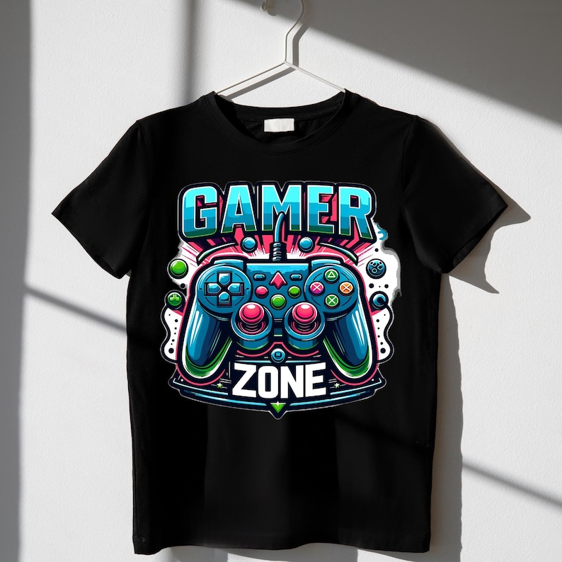 May include: Black t-shirt with a colourful graphic of a video game controller and the words "GAMER ZONE". The controller is blue, red, and green, with a detailed illustration. The t-shirt is hanging on a white hanger.
