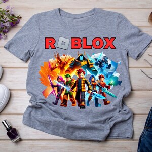 May include: Grey t-shirt with the word "ROBLOX" in red and a grey square. Below the text is a colourful graphic of characters with weapons and special effects. The shirt is made of a soft material.