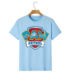 May include: A light blue t-shirt featuring the "PAW PATROL" logo. The logo is a shield shape with a red background, blue and yellow lettering, and a white paw print. The shirt is hanging on a wooden hanger.