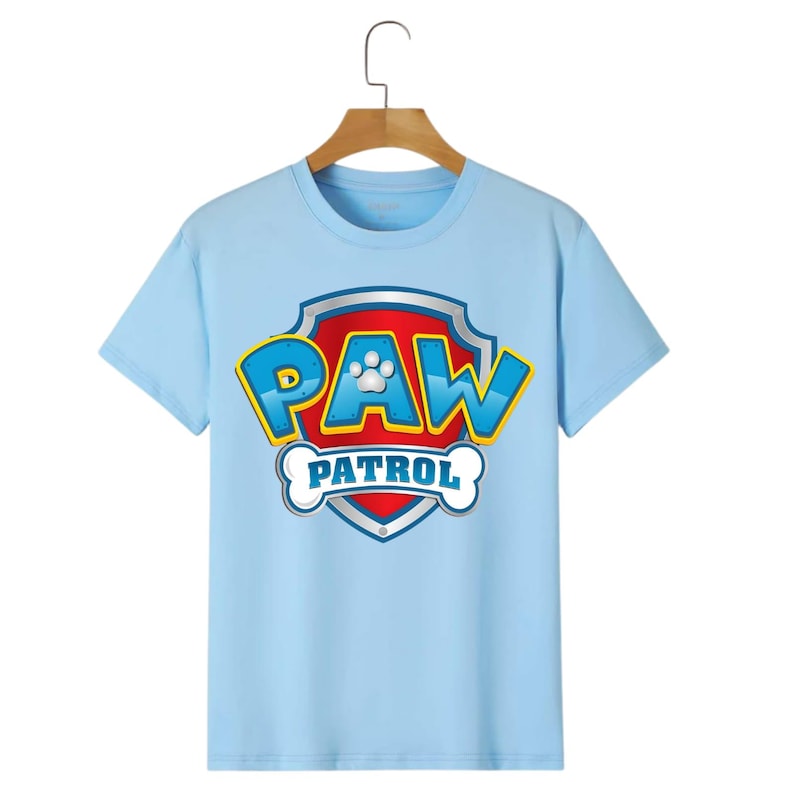 May include: A light blue t-shirt featuring the "PAW PATROL" logo. The logo is a shield shape with a red background, blue and yellow lettering, and a white paw print. The shirt is hanging on a wooden hanger.