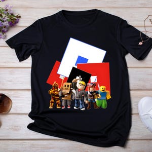 May include: Black t-shirt with a Roblox graphic. The design shows several Roblox characters in various outfits, set against a backdrop of red, white, blue, and black geometric shapes. A fun and colourful design.