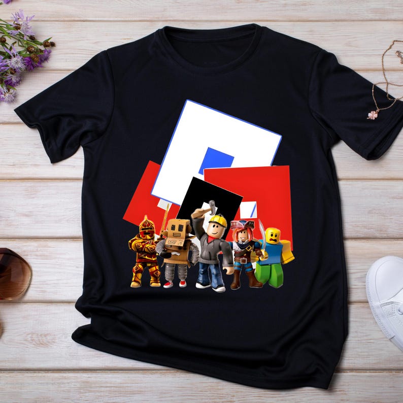 May include: Black t-shirt with a Roblox graphic. The design shows several Roblox characters in various outfits, set against a backdrop of red, white, blue, and black geometric shapes. A fun and colourful design.
