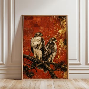 以下が含まれることがあります： 枝に止まっている2羽の鷹が描かれた額入りのアートプリント。背景は金色のアクセントが入った質感のある赤色。鳥は茶色と白で詳細に描かれており、鮮やかなコントラストを生み出しています。