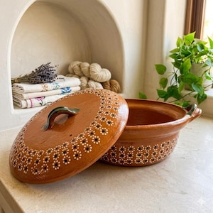 May include: A terracotta-colored ceramic casserole dish with a matching lid. The lid and the rim of the dish are decorated with a pattern of small white and black floral designs. The handle is green.