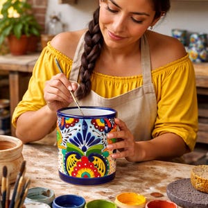 May include: A woman paints a ceramic vase with a colorful floral design. The vase is cylindrical with a blue base and rim. Various paint colors in small containers and paintbrushes are on the table. The woman wears a yellow top and a beige apron.