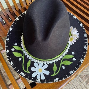May include: A dark brown fedora hat with a decorative brim. The brim is black with white daisy and green leaf accents. A white daisy and green leaves are on the side of the hat. A green band encircles the crown.
