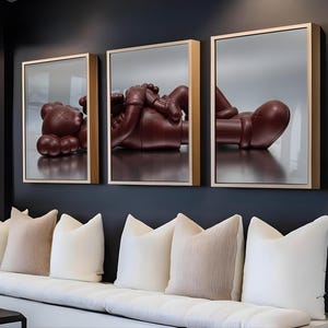 Pode incluir: Três obras de arte emolduradas com uma escultura marrom de uma figura com olhos em forma de X e corpo arredondado. A obra de arte é exibida acima de um banco branco com várias almofadas em uma sala de estar moderna.