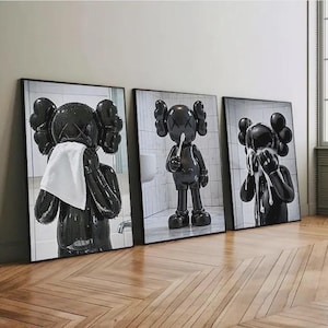 May include: Three framed art prints featuring a black KAWS figure in a bathroom setting. One figure holds a towel, another brushes its teeth, and the third washes its face. The prints have black frames and are displayed on a light-colored wall.