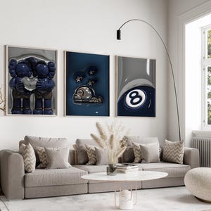 May include: A modern living room featuring three framed art pieces above a gray sectional sofa. The artwork includes a blue figure holding a heart, a dark blue abstract piece, and a close-up of a pool ball. A white coffee table and a curved floor lamp complete the scene.