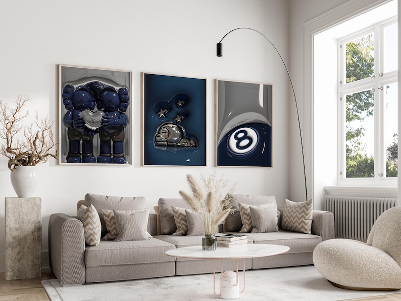 May include: A modern living room featuring three framed art pieces above a gray sectional sofa. The artwork includes a blue figure holding a heart, a dark blue abstract piece, and a close-up of a pool ball. A white coffee table and a curved floor lamp complete the scene.