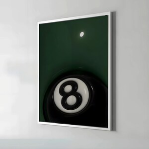 May include: A framed print featuring a close-up of a black and white eight ball. The eight ball is set against a dark green background, with a bright white circle in the upper portion of the image. The print is encased in a white frame.