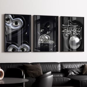 Op de afbeelding: Drie ingelijste kunstdrukken met zwarte frames. De prints tonen abstracte, metallic ontwerpen in zwart en zilver. Eén print toont ogen, een andere een metalen object met sterren en de derde een poolbal met het Stussy-logo.