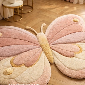 K&ouml;nnte beinhalten: Ein gro&szlig;er, pl&uuml;schiger Schmetterlings-Teppich in Rosa-, Creme- und Gelbt&ouml;nen. Der Teppich hat ein detailliertes Design mit strukturierten Fl&uuml;geln und einem zentralen K&ouml;rper. Der Text "AaradhyaCarpetArtt" ist unten auf dem Teppich zu sehen.